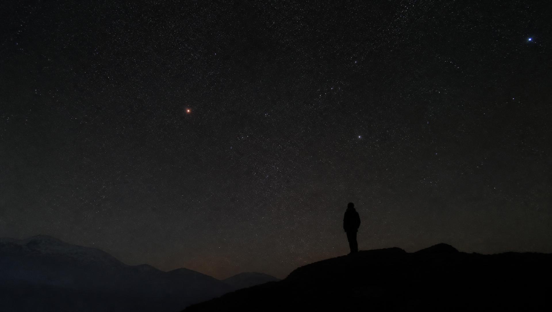 A lone figure on a dark ridge beneath a dense field of stars, with Mars visible as a small red point of light