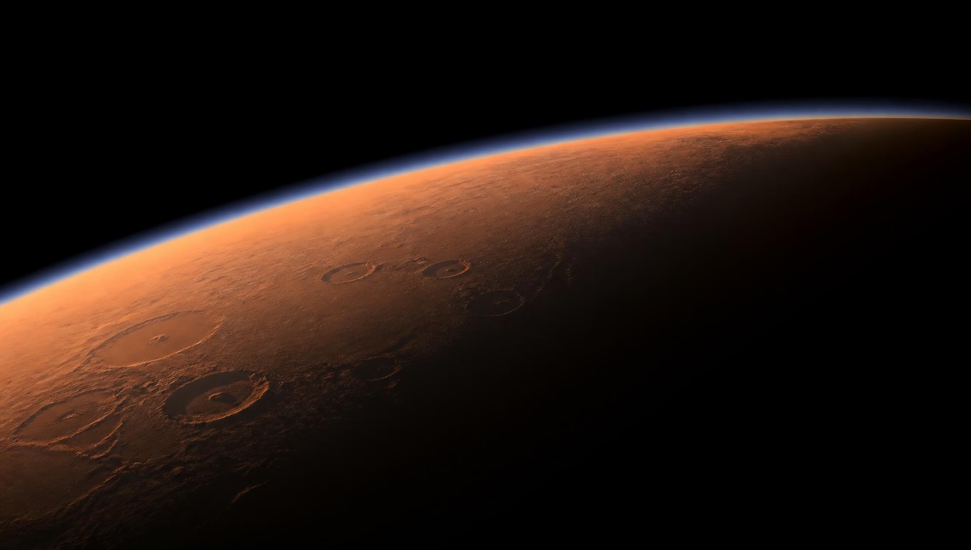 Mars seen from orbit at the terminator line, a thin bright atmosphere arcing over rust-coloured cratered terrain